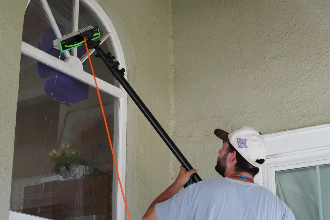 Close view of a technician angling the water fed pole brush onto an arched window beside a door, orange hose along the pole