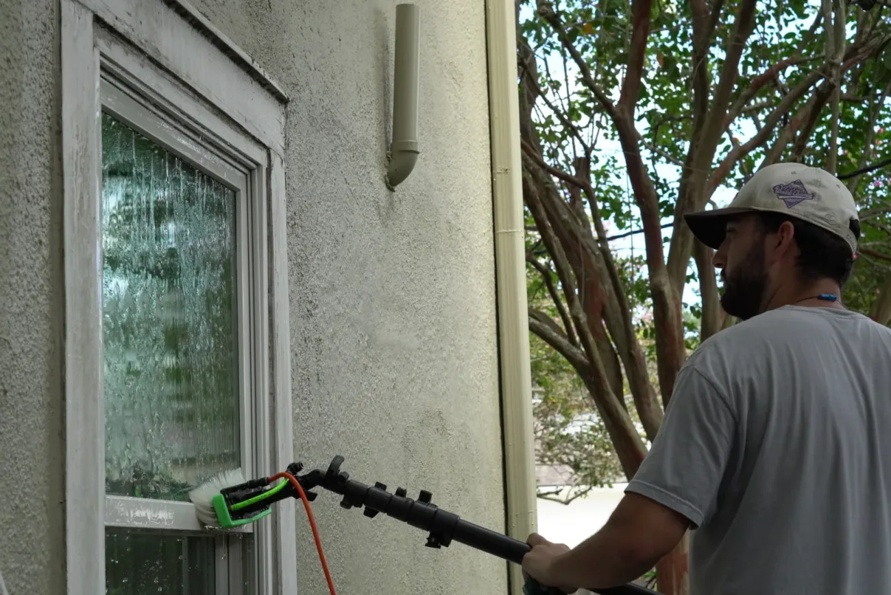 Boot Krewe Cleaners technician running the pure-water brush across a window on a cream stucco wall, water sheeting down the glass