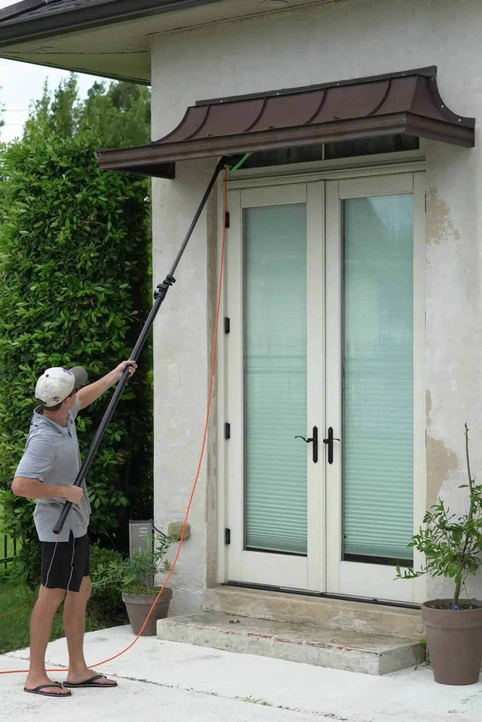 Technician in a cap running a water fed pole up over the copper awning above cream french doors, orange hose trailing