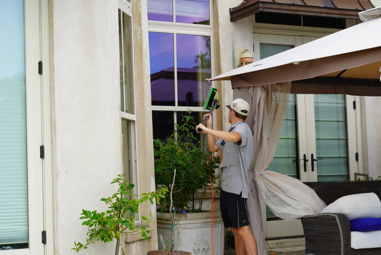 Boot Krewe Cleaners technician reaching up with a water-fed pole to a tall window beside a patio umbrella on a cream stucco house