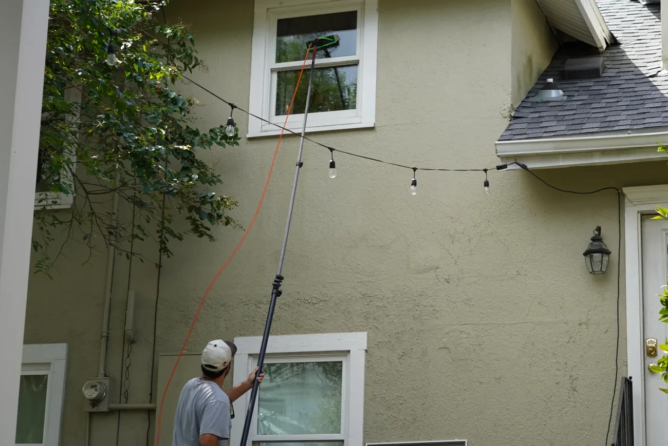 Technician on the ground running a water-fed pole with an orange hose up to an upstairs window on a green stucco house